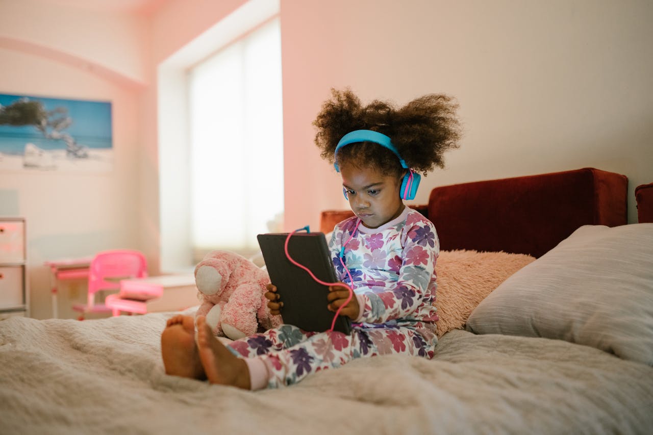 A young girl in pajamas sits on the bed with headphones, using a tablet. Cozy home setting.