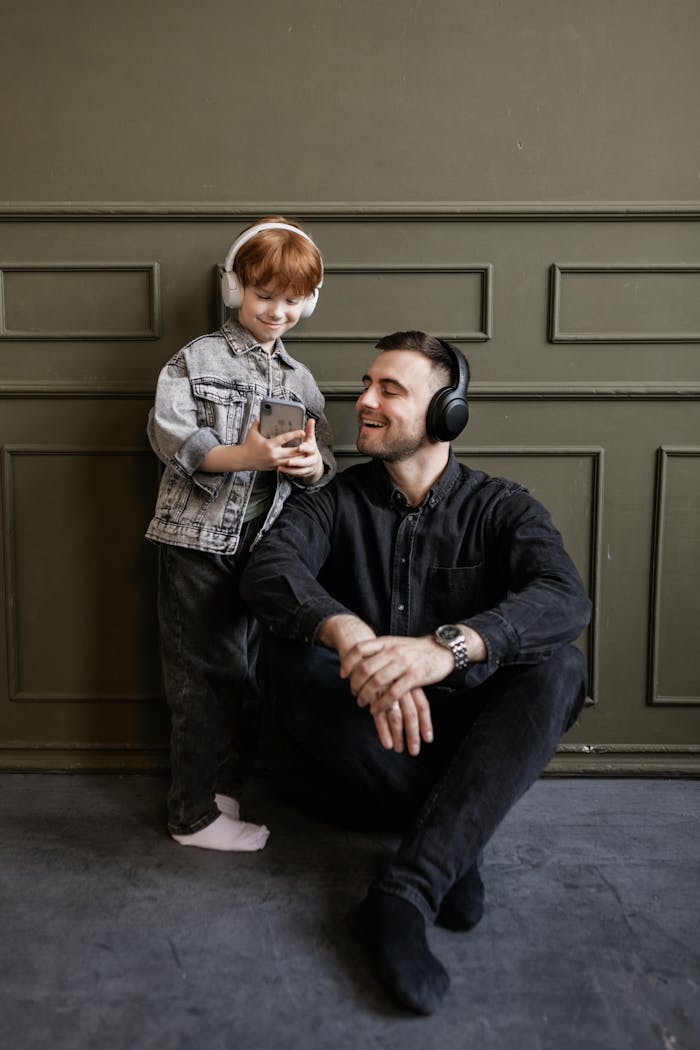 Father and son smiling while listening to music on headphones indoors.